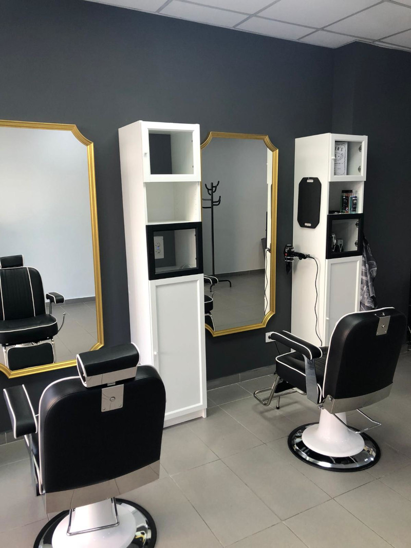 Otra vista del interior de la peluquería mostrando dos estaciones de trabajo adicionales. Las estaciones también tienen espejos con marcos dorados y estanterías blancas. Las sillas de barbero son similares, de color negro con detalles en blanco. La peluquería tiene un estilo moderno y elegante.