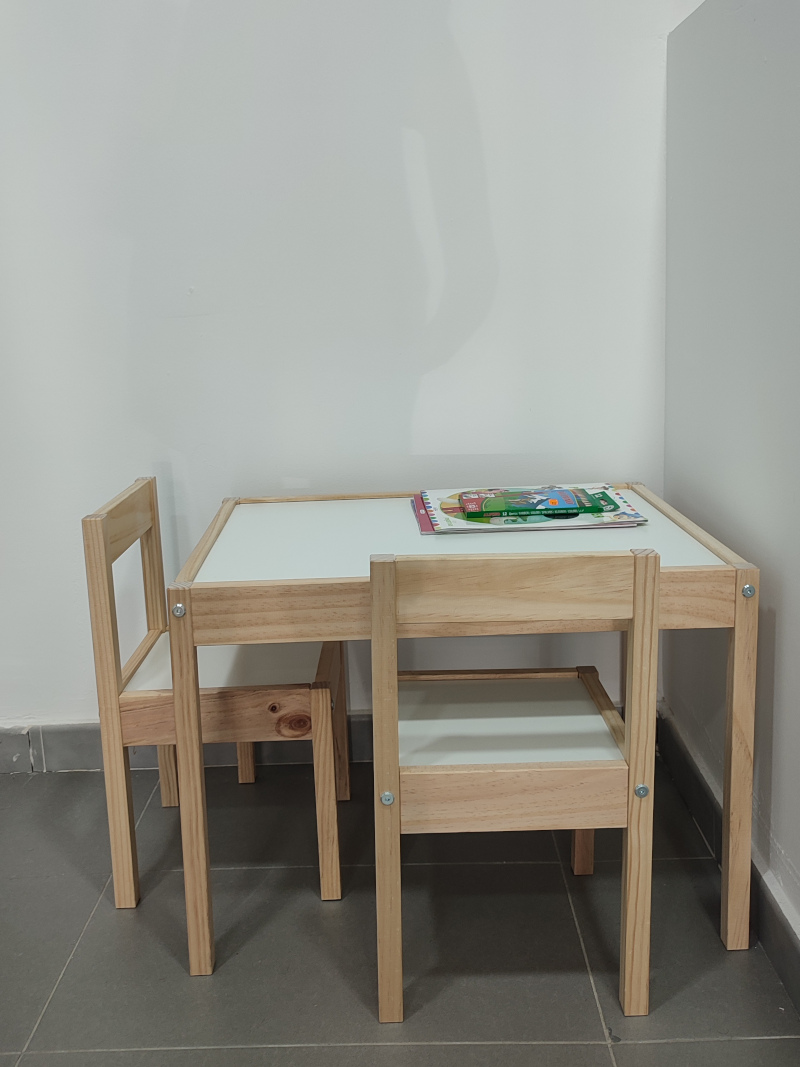 Interior de una peluquería, Pequeño escritorio de madera con dos sillas para niños, colocado contra una pared blanca. Sobre el escritorio hay algunos libros y lápices de colores. El conjunto está en una esquina bien iluminada.