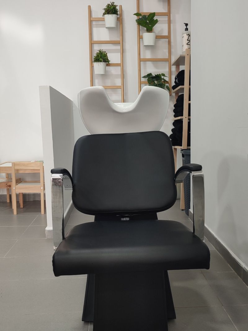 Interior de una peluquería, se muestra un área de lavado con un sillón de lavado negro, con respaldo y asiento acolchados y reposabrazos cromados, colocado frente a un lavacabezas blanco. Detrás del sillón, hay dos escaleras decorativas de madera clara, cada una con varias macetas blancas colgantes que contienen plantas verdes. A la derecha de las escaleras, hay un estante de madera con toallas negras dobladas, y una botella grande de champú con dispensador se encuentra en la parte superior del estante.