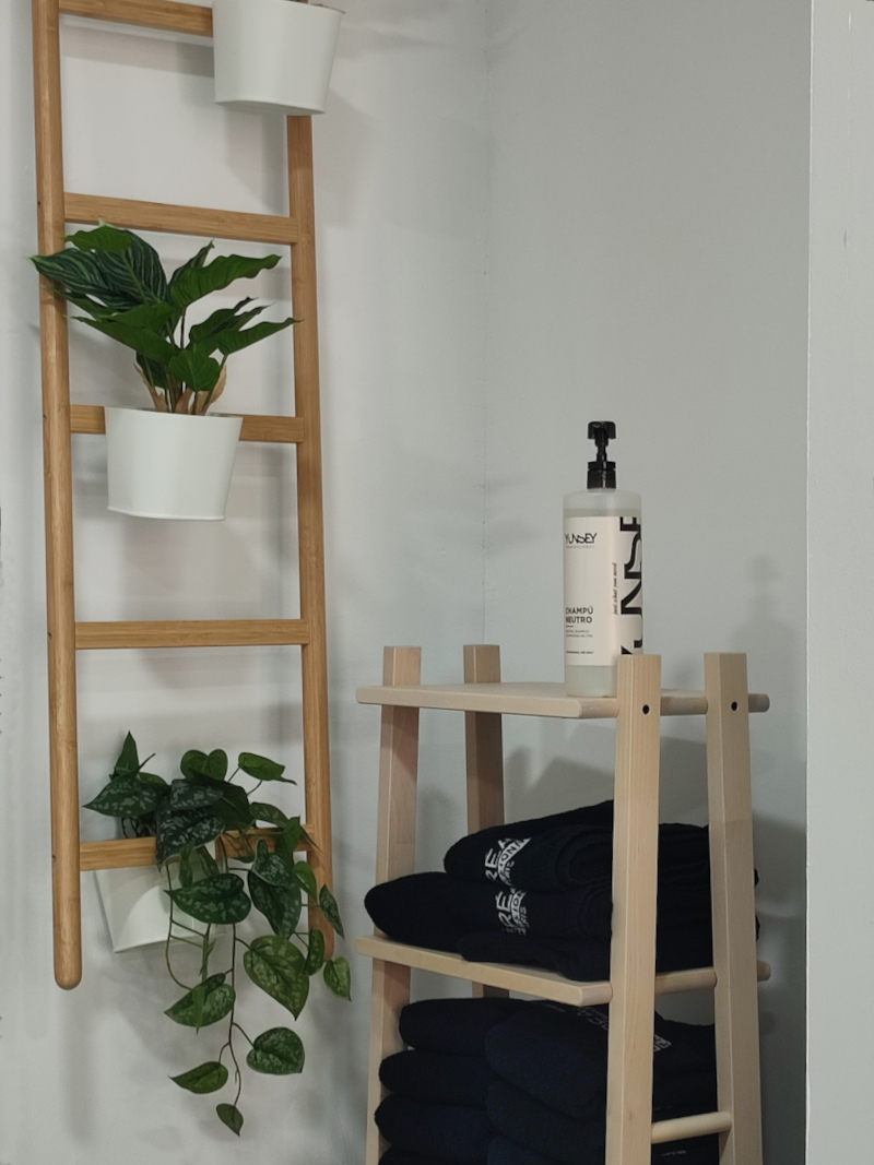 Interior de una peluquería con una esquina decorativa. En la pared se encuentra una escalera decorativa de madera clara con tres macetas blancas colgantes, cada una con una planta verde. Frente a la escalera, hay un pequeño estante de madera clara con varios estantes llenos de toallas negras dobladas. En la parte superior del estante hay una botella grande de champú con dispensador.